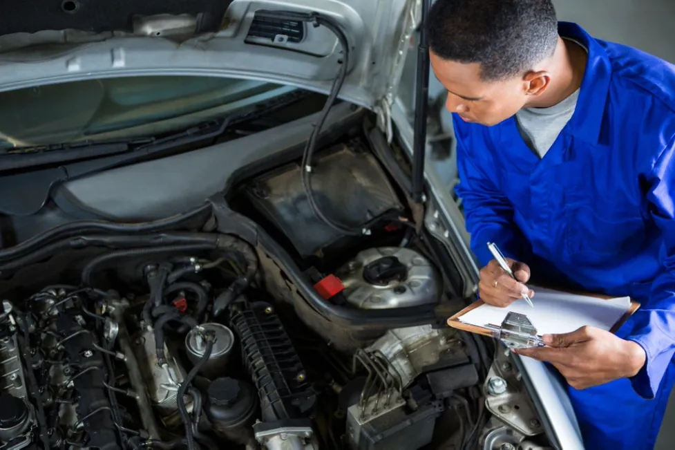 A mechanic in a blue jumpsuit carefully inspects a car engine while taking notes on a clipboard.
