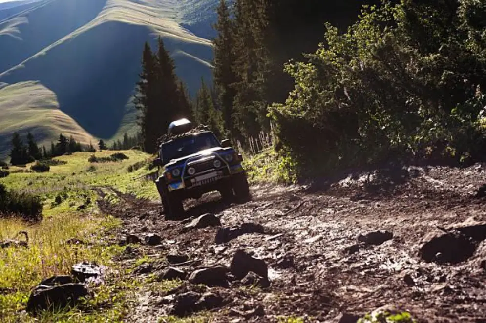 A 4x4 vehicle, likely a Jeep Wrangler, navigating a muddy, rocky uphill trail in a natural, mountainous, and forested landscape under a bright sky.
