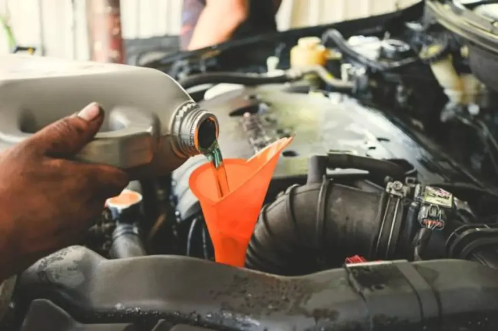 A person's hands pouring fresh fluid from a bottle, using an orange funnel, into an automotive fluid reservoir under the hood of a vehicle. The engine bay is visible.