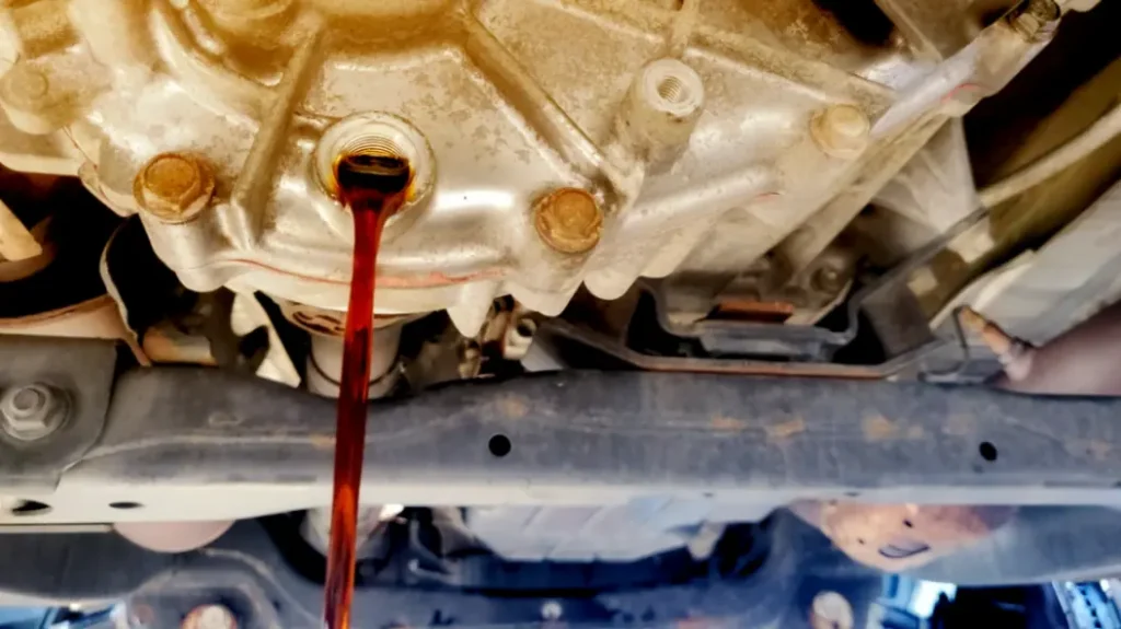 A reddish-brown fluid is being drained from the underside of a vehicle, indicating a fluid change in progress. The vehicle's undercarriage and part of the tire are visible.