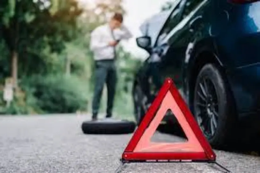 A person standing next to the front of a car with a red warning triangle on the ground in front of the vehicle, indicating a breakdown or roadside issue.