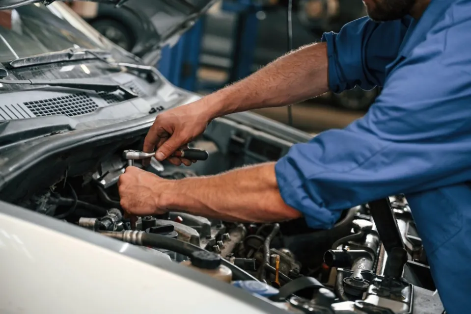 A mechanic's hands are visible, performing maintenance work under the hood of a car, with various engine components around.